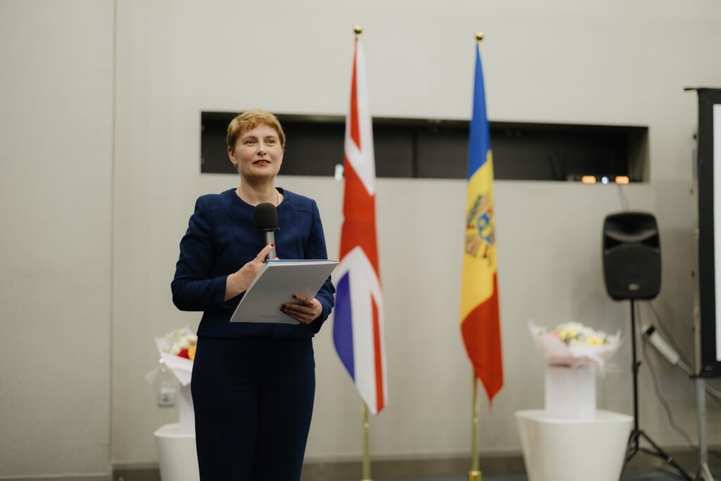 Dina Railean, Honorary Consul of the Republic of Moldova in Manchester, delivers a formal speech at an official diplomatic event. She stands holding a microphone and notes, positioned between the national flags of the United Kingdom and the Republic of Moldova. Wearing a navy-blue business suit, she addresses guests in a modern reception venue. Floral arrangements and presentation equipment are visible in the background, reflecting the formal nature of the occasion. The image symbolises diplomatic relations, UK–Moldova cooperation, community leadership, international partnership and the strengthening of ties between Moldova and the North West of England.