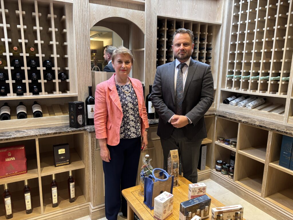 Dina Railean, Honorary Consul of Moldova in Manchester in a coral blazer and Alexandru Vlasenco in a dark suit stand inside the Moldovan Embassy’s wine vault in London, surrounded by wooden shelves displaying Moldovan wines, spirits and traditional products. The photograph followed a discussion about business and investment opportunities in Moldova, the role of the Honorary Consulate in promoting these opportunities across the North West of England, and future engagement with the Moldovan diaspora.