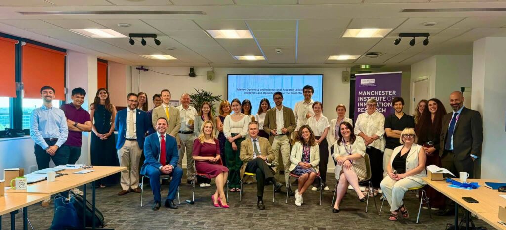 Group photograph of diplomats, academics, researchers and regional stakeholders attending the “Science Diplomacy and International Research Collaboration in the North West” seminar at Alliance Manchester Business School, University of Manchester, on 9 July 2026. Attendees pose in a conference room beside a Manchester Institute of Innovation Research banner, with the event title displayed on a screen behind them. The seminar explored place-based science diplomacy, European research collaboration and stronger links between universities, industry, public authorities and the diplomatic community.
